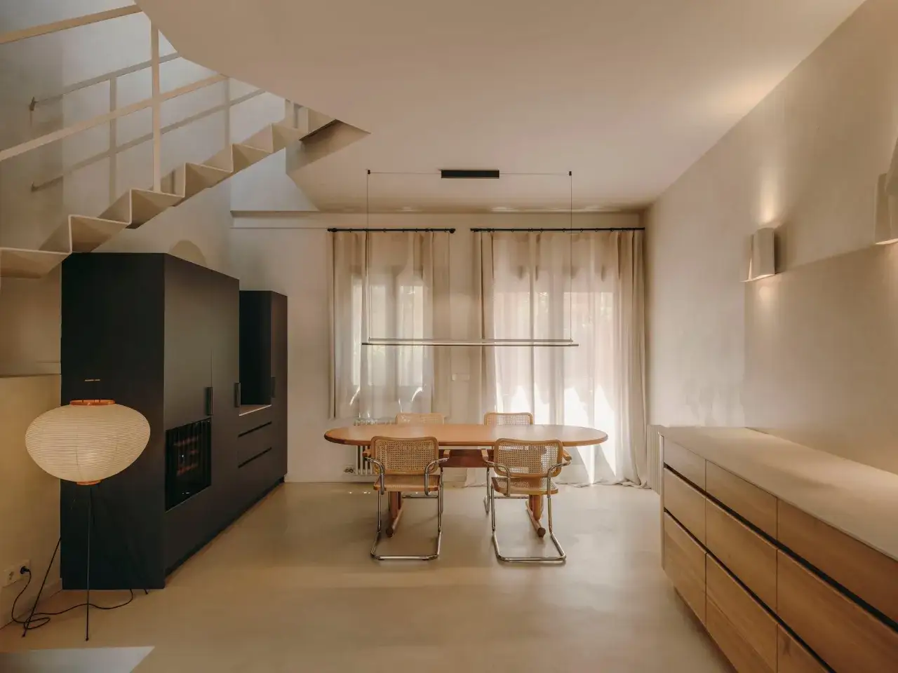 Comedor moderno con suelo de **b&eacute;ton cir&eacute;**, mesa de madera, sillas de mimbre y una escalera flotante.