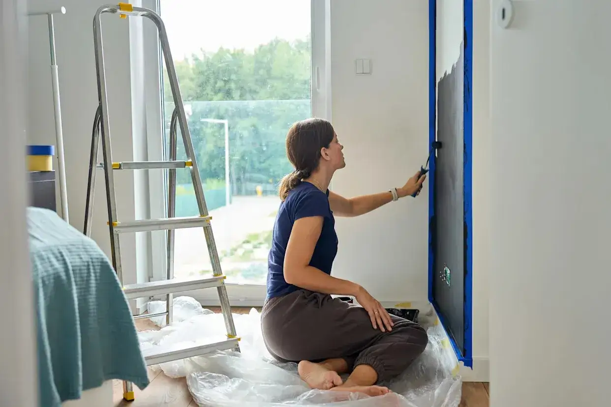 Frau malt mit einer Rolle eine Wand schwarz an. Sie sitzt auf dem Boden, um den Lack auf Lack zu streichen. Eine Leiter steht daneben.