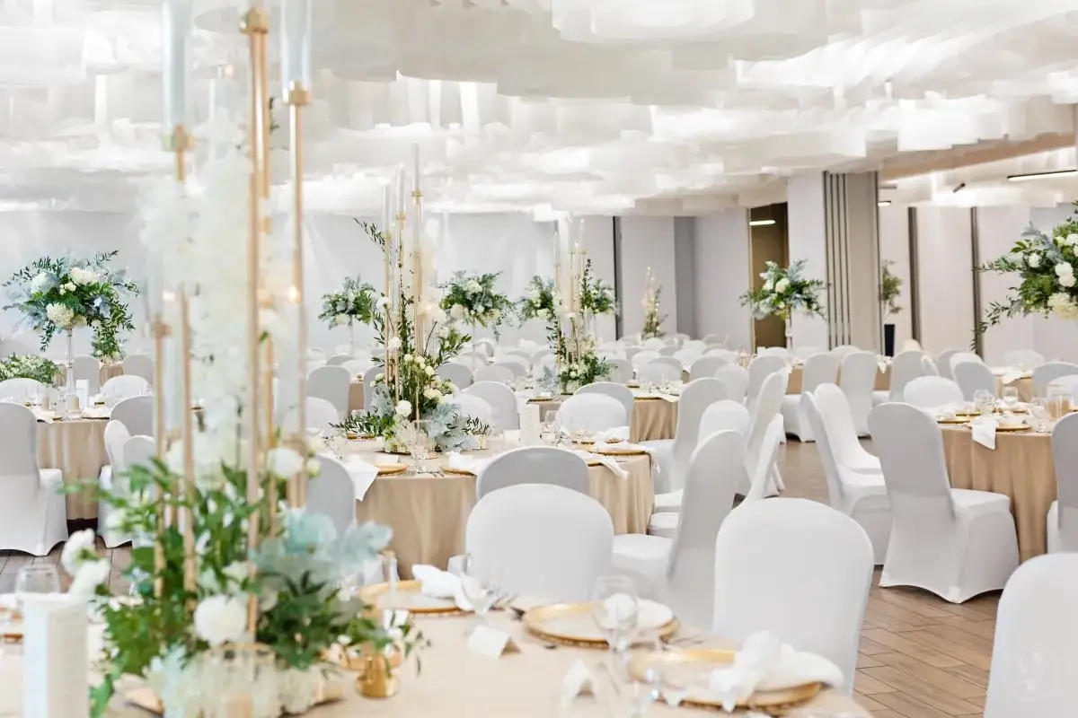 Elegancka sala bankietowa hotelu z białymi pokrowcami na krzesłach, złotymi zastawami i bukietami kwiatów. Idealne miejsce na przyjęcie z **luxury collection hotel**.