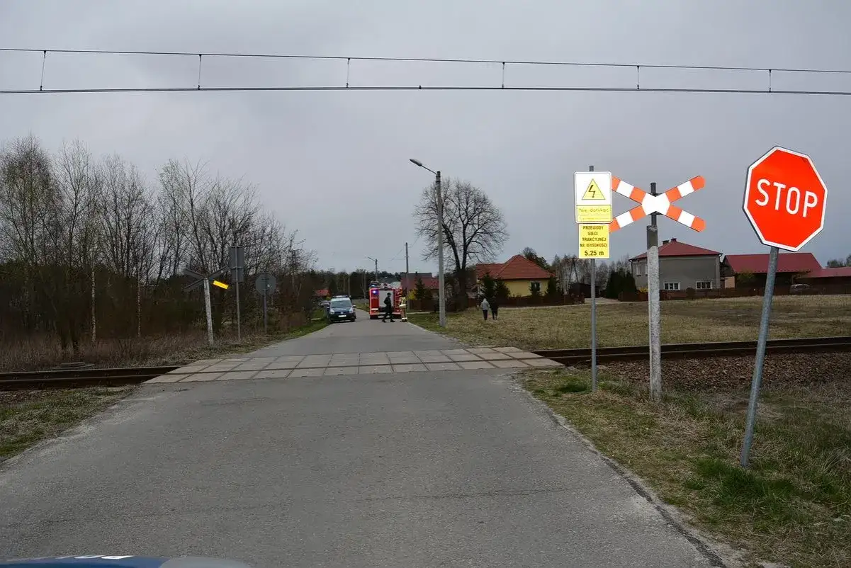 Przejazd kolejowy z znakiem G3, krzyżem św. Andrzeja, znakiem STOP i ostrzeżeniem o przewodach trakcyjnych. W oddali radiowóz i wóz strażacki.