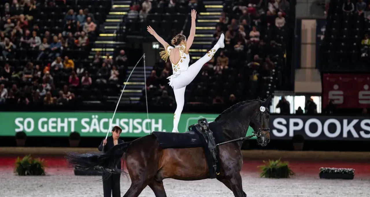 Eine Voltigiererin in weiß-goldener Kleidung steht auf einem Pferd, bereit für das Voltigiieren Olympia.