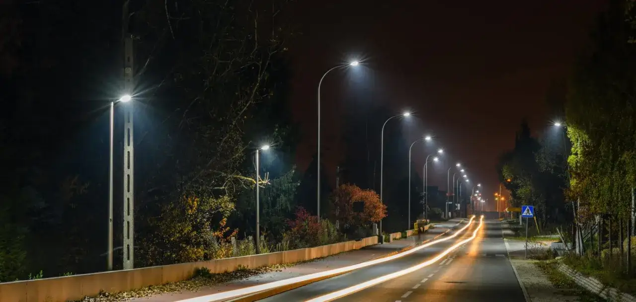 Droga nocą, rozświetlona latarniami. Długie światła samochodów tworzą smugi na asfalcie. Jak załatwić oświetlenie ulicy? Tutaj widać przykład.