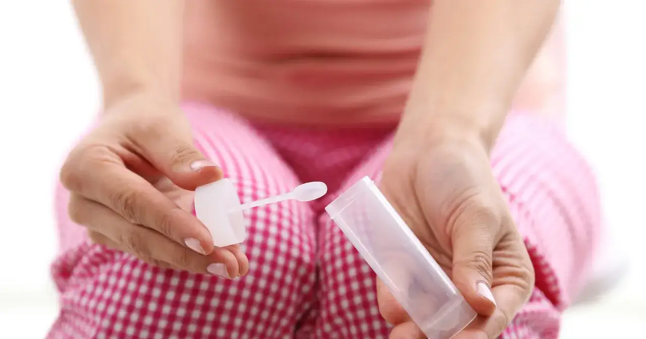 Dłoń trzyma pojemnik z łyżeczką do pobierania próbek kału, potrzebny do badania poziomu kalprotektyny w kale.