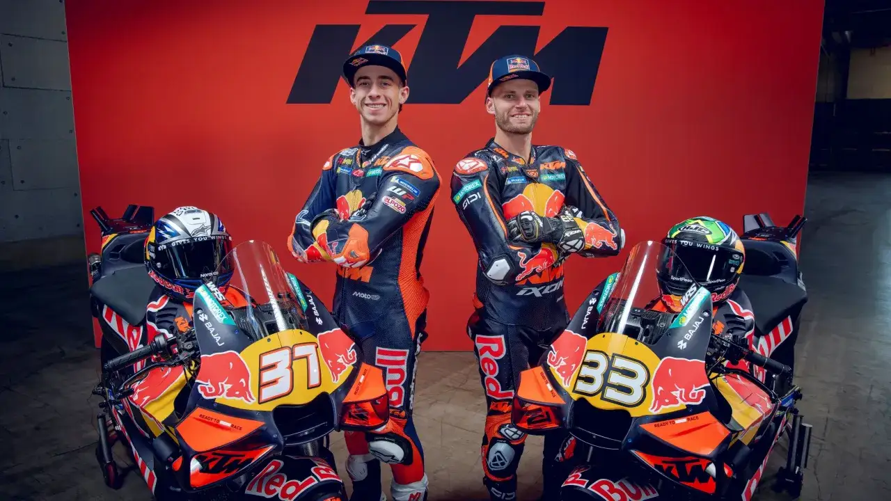 Dos pilotos de MotoGP KTM posan con sus motos. Los pilotos visten trajes de carreras naranjas y negros, y las motos son de color naranja y negro con el logo de Red Bull.