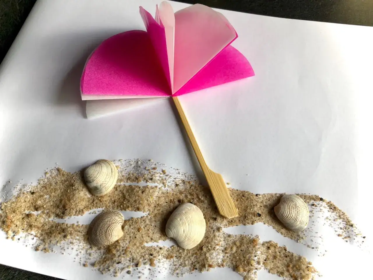 Ein selbstgebastelter Sonnenschirm aus Papier und Holzspie&szlig; steht auf Sand mit Muscheln. Perfekt f&uuml;r eine Bastelidee zum Thema Sommer.