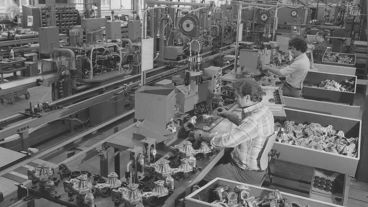 Dans une usine, des ouvriers assemblent des pièces. On voit des machines et des boîtes remplies de composants, typiques de ce qui fabrique la marque Bosch.