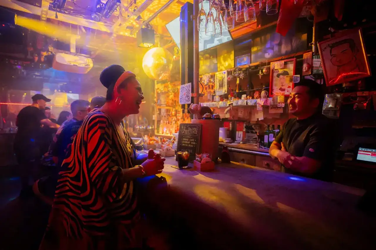 A vibrant scene inside a gay sauna in Osaka. A person in a striking zebra-print top chats with a bartender behind a well-stocked bar, bathed in colorful, smoky light.