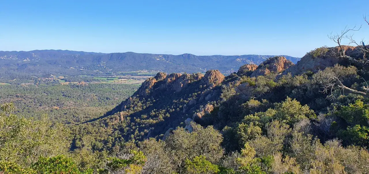 Paisaje monta&ntilde;oso de las Gavarres con rocas rojizas y vegetaci&oacute;n frondosa bajo un cielo azul despejado.
