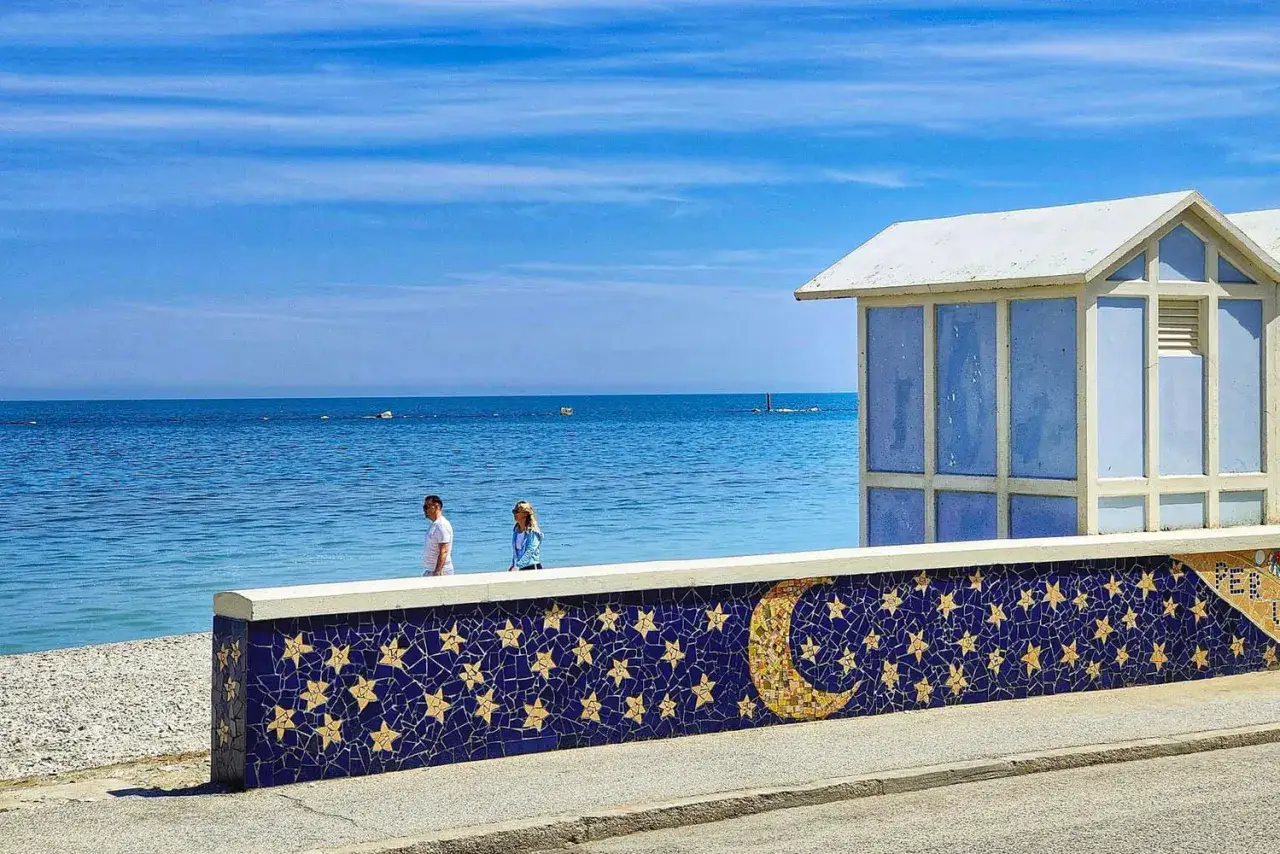 Una coppia passeggia sulla spiaggia marotta, con un muretto decorato con stelle e luna che separa la sabbia dal mare azzurro.