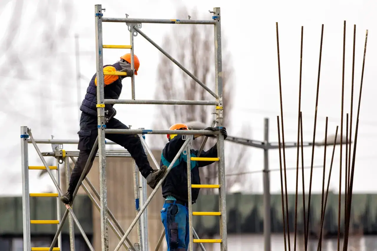Dwóch budowlańców w kaskach pracuje na rusztowaniu, przestrzegając zasad BHP.