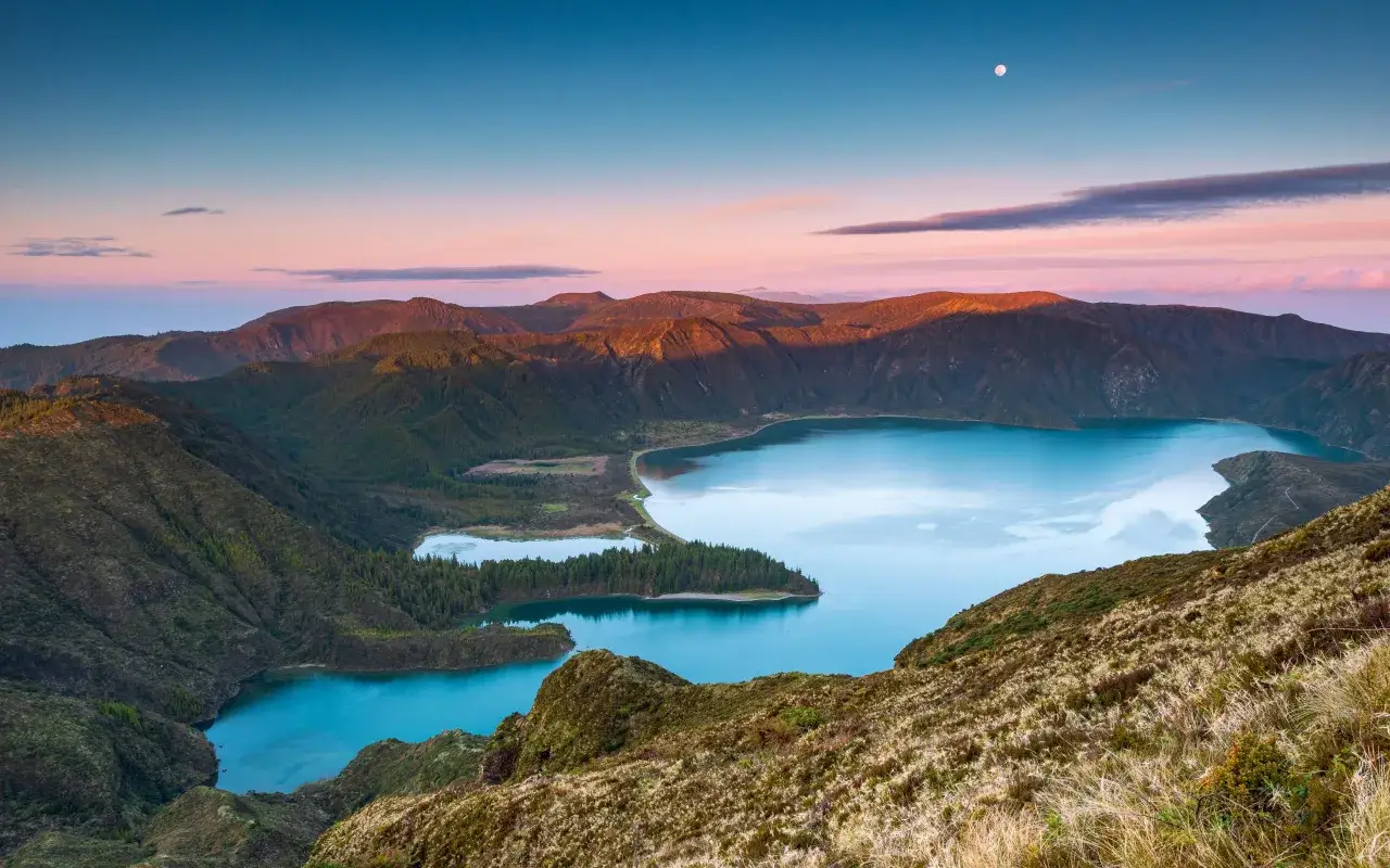 Paisaje al amanecer en Lagoa do Fogo, Azores. Monta&ntilde;as verdes rodean lagos de azul intenso bajo un cielo rosado y una luna p&aacute;lida.