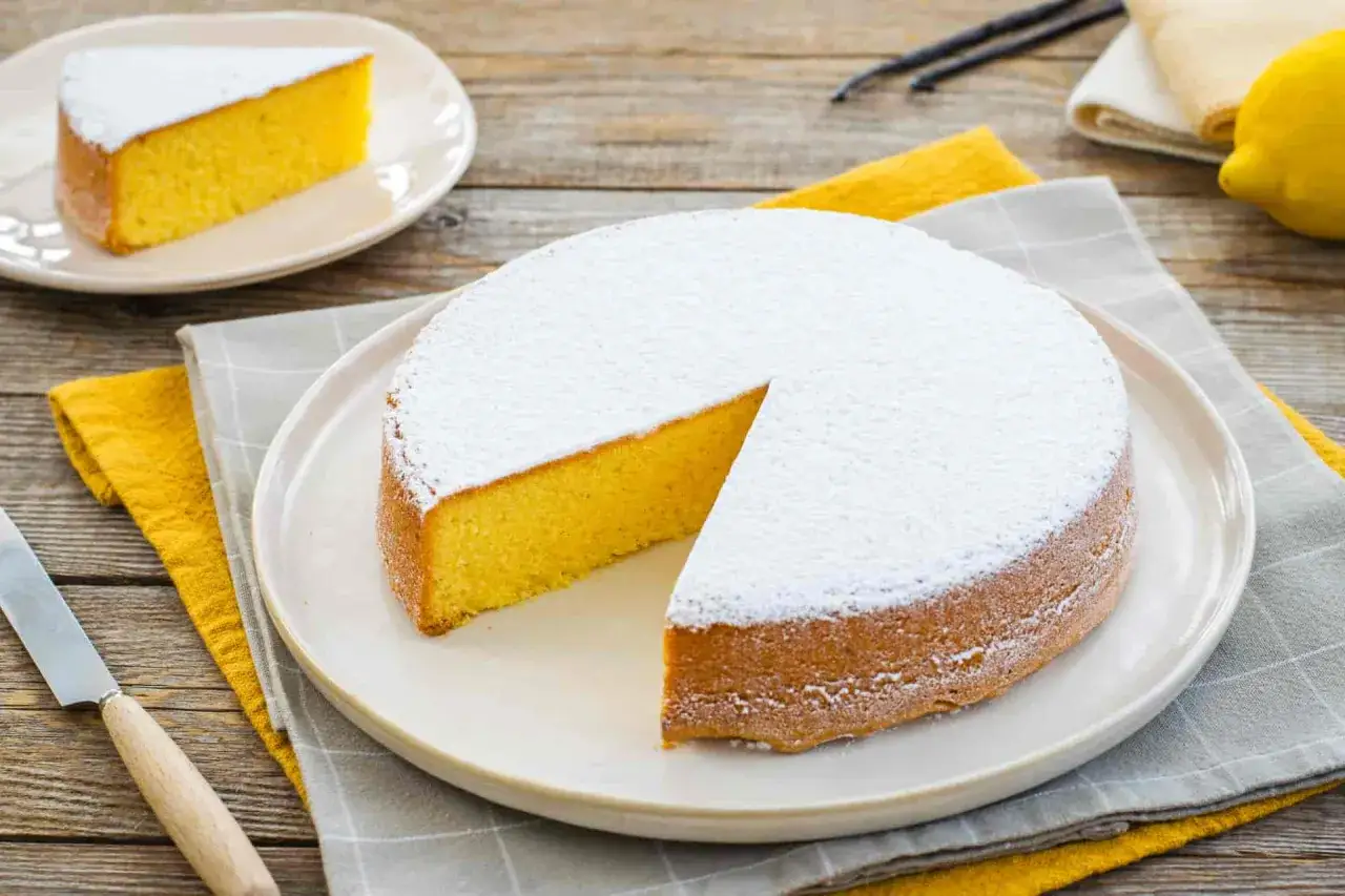 Una torta paradiso sofficissima, cosparsa di zucchero a velo, con una fetta già servita su un piattino. Ingredienti freschi come limone e vaniglia completano la scena.