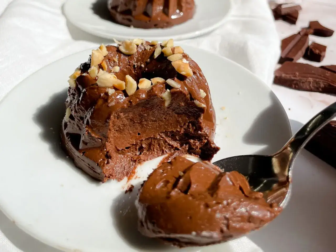 Budino fatto in casa al cioccolato, cremoso e ricco, guarnito con nocciole tritate. Un cucchiaino ne preleva una porzione golosa.