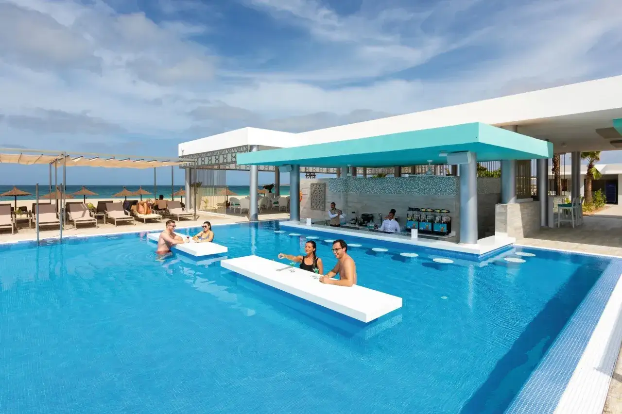 Paare genießen Drinks an der Swim-up-Bar im Riu Palace Boavista. Sonnenschirme und Liegen am Strand im Hintergrund.