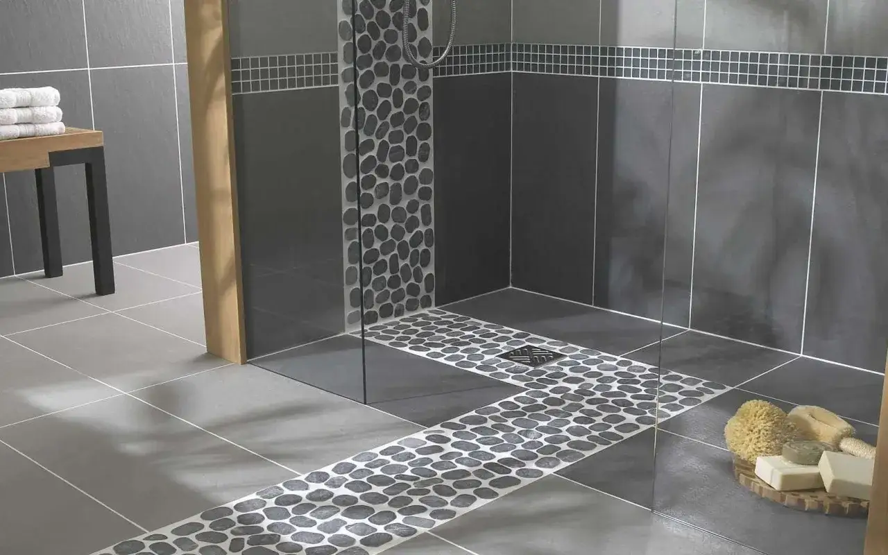 Douche italienne moderne avec carrelage imitation pierre de galets sur le sol et le mur. Un banc en bois et des serviettes blanches complètent l'ensemble.