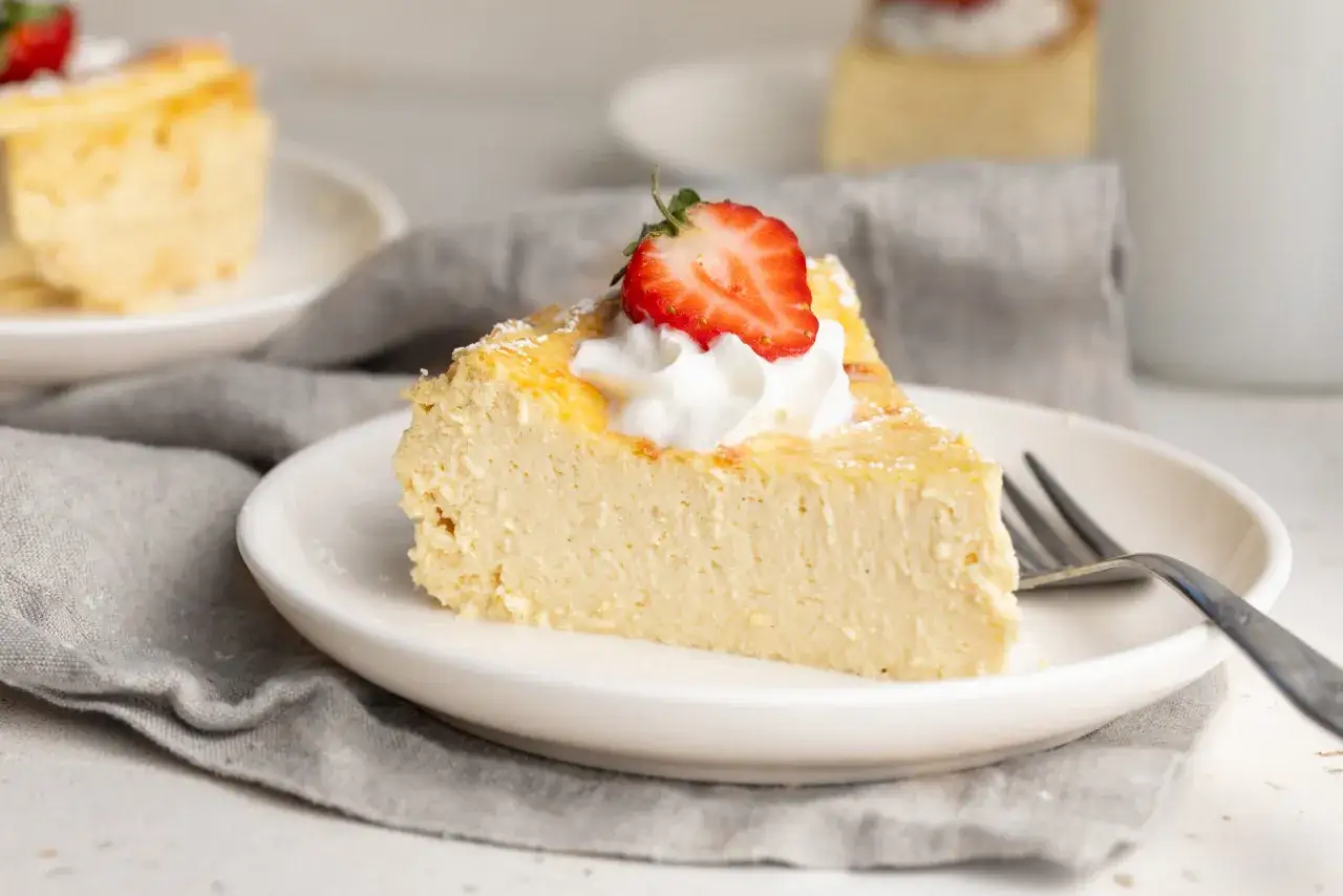 Rebanada de tarta de queso light cremosa, decorada con nata y fresa, servida en plato blanco con tenedor.