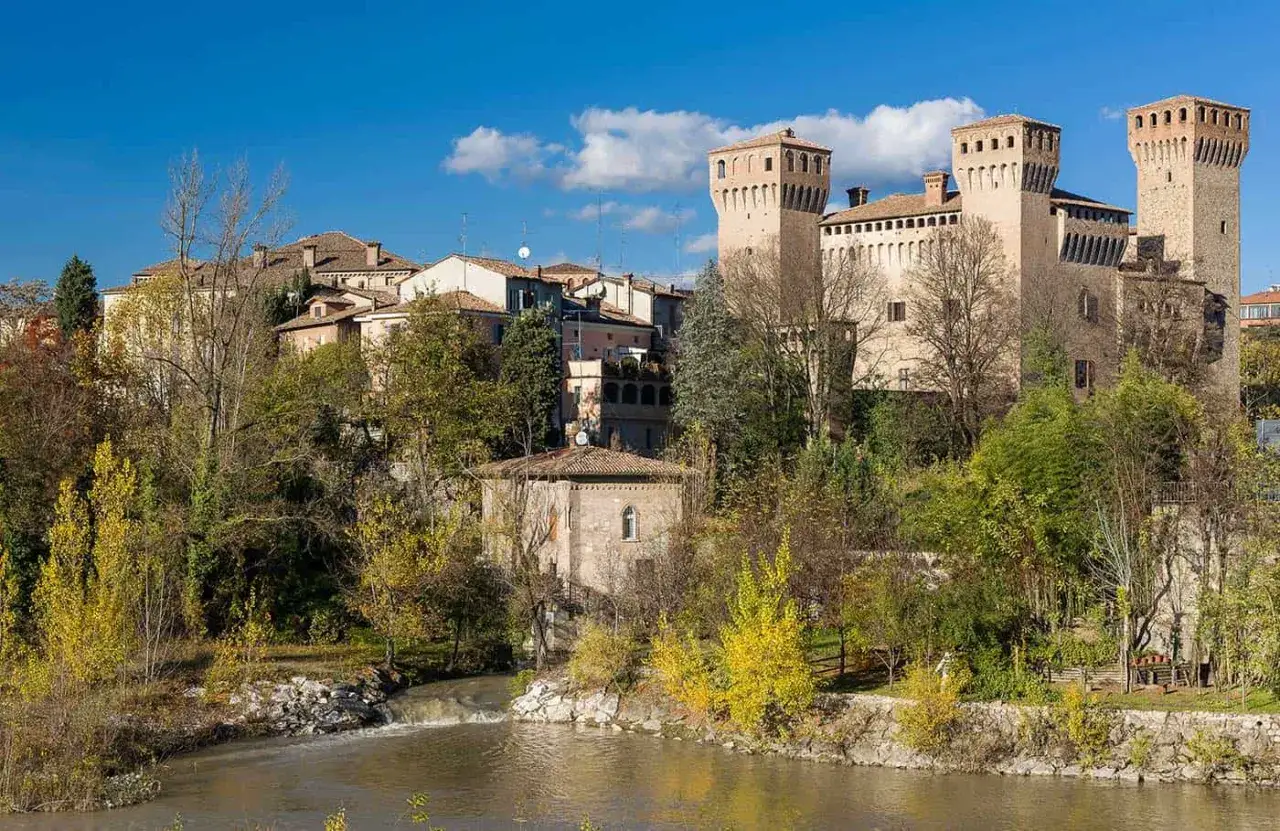 Castello medievale lungo la strada romea, con torri imponenti e un fiume che scorre placido tra alberi autunnali.