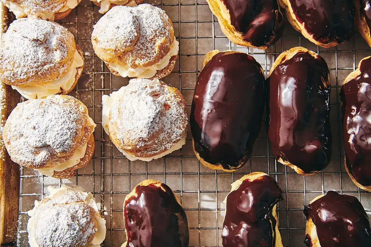 Puszyste ptysie z kremem patissiere i cukrem pudrem obok błyszczących czekoladą eklerów.