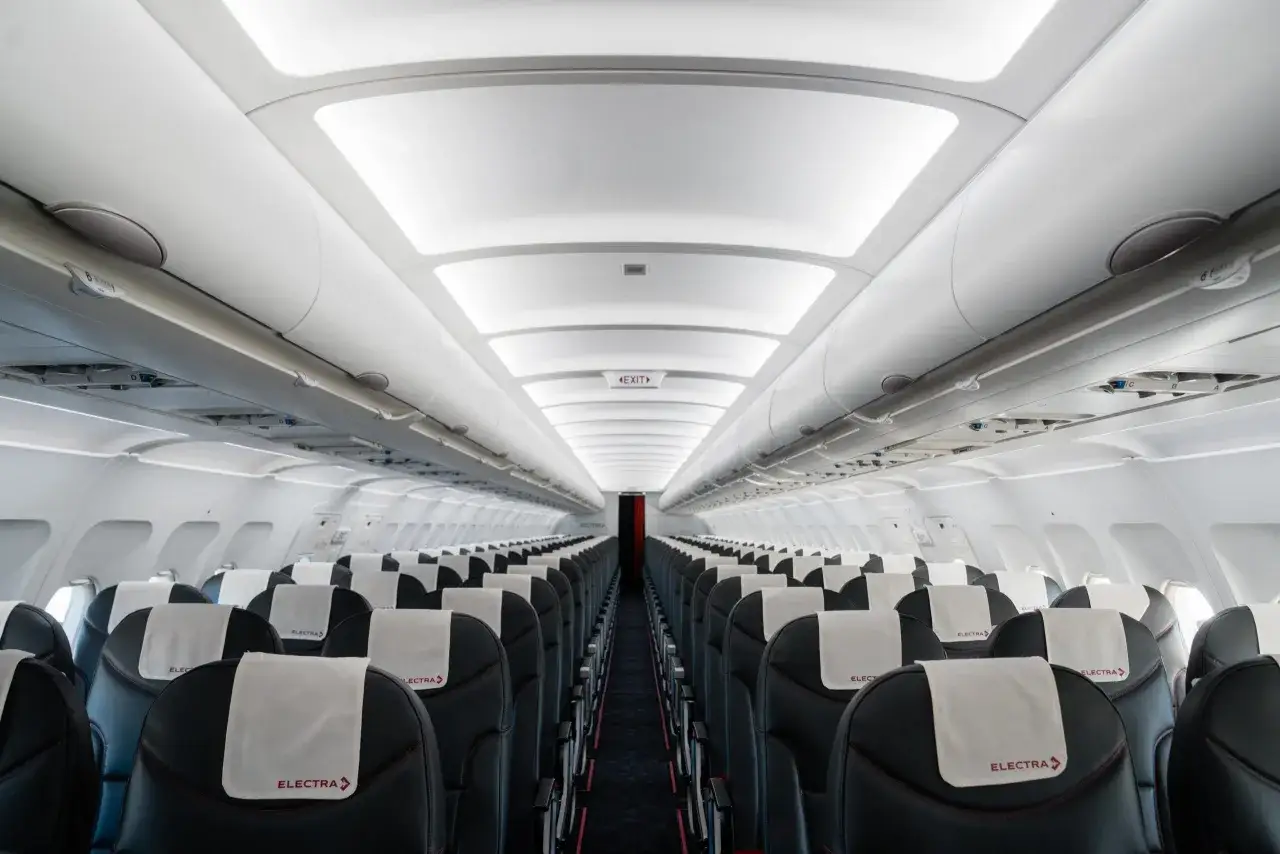Wnętrze samolotu linii lotniczych Electra Airways z rzędami czarnych foteli i białymi zagłówkami z logo.
