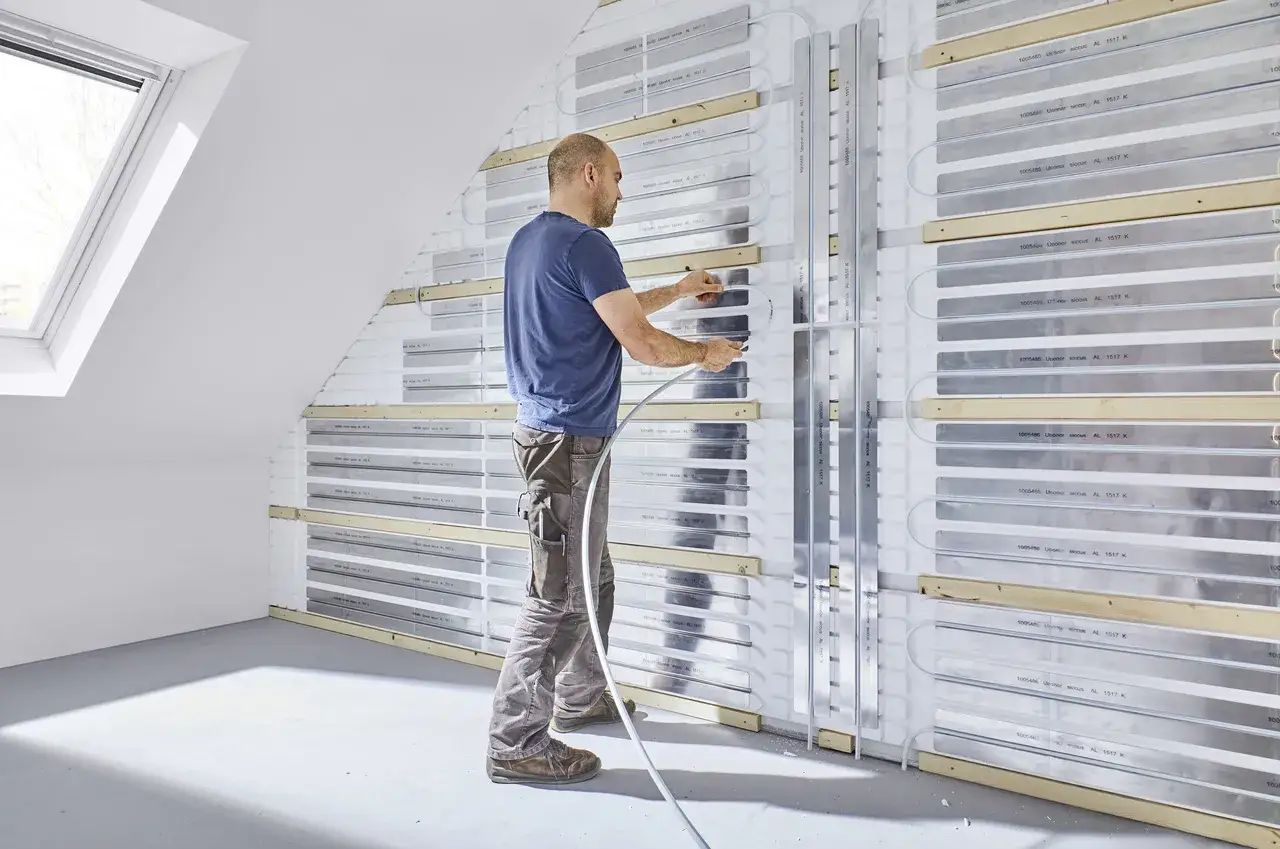 Instalator montuje system ogrzewania ściennego wodnego, układając rurki w specjalnych aluminiowych profilach.