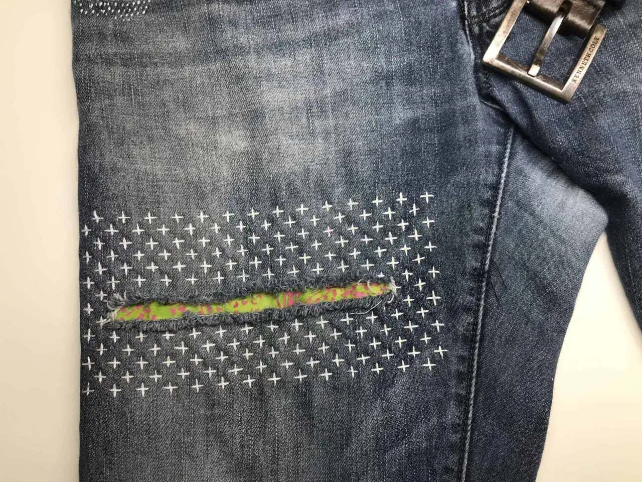 A close-up of denim jeans showcasing a creative sashiko jeans repair. A colorful fabric patch peeks through a tear, mended with white cross-stitch embroidery.