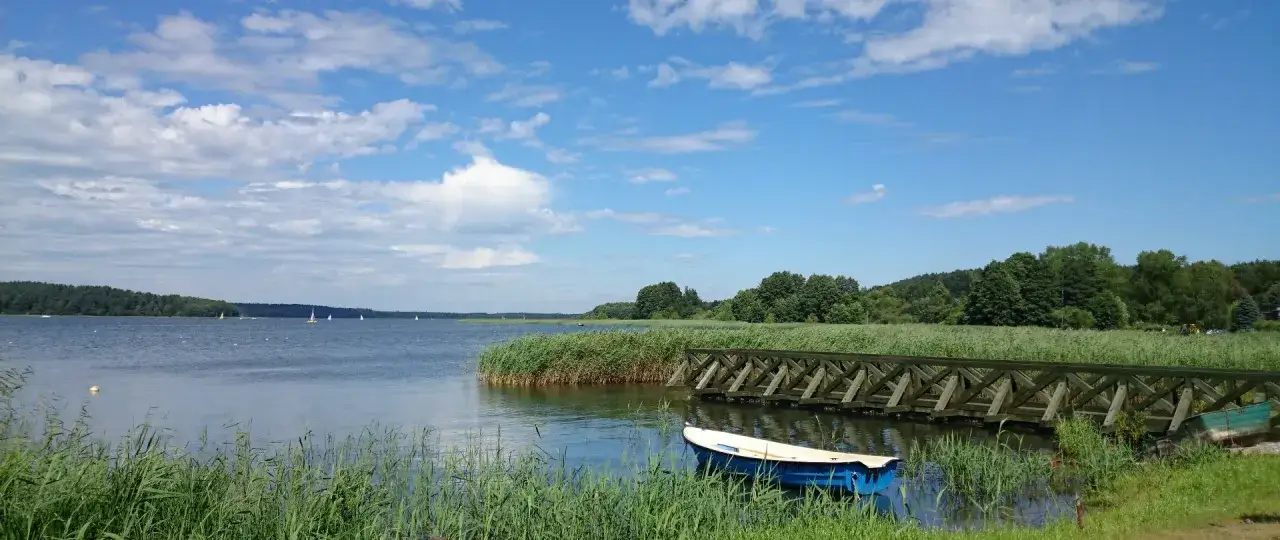 Błękitne niebo z chmurami nad jeziorem, gdzie są Bory Tucholskie. Drewniany pomost, ł&oacute;dka i żagl&oacute;wki na wodzie.