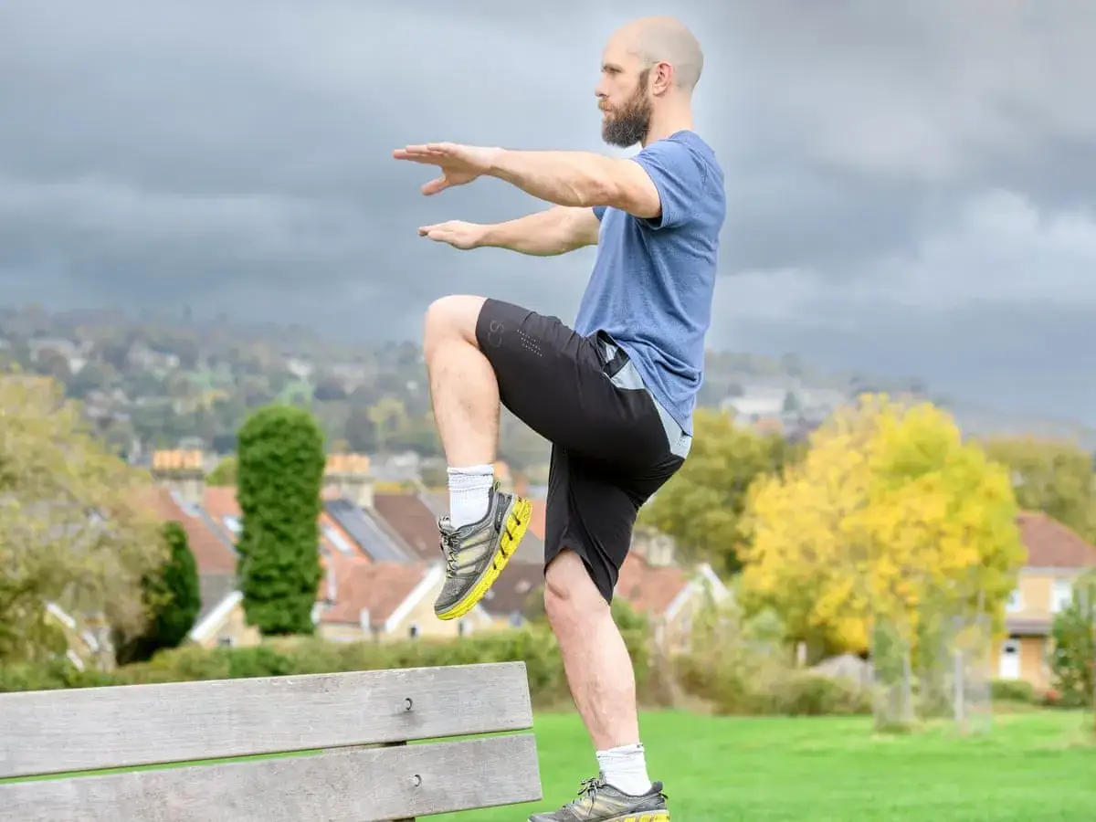 Ein Mann mit Glatze und Bart steht auf einem Bein auf einer Parkbank. Er trainiert seine Balance, während er auf einem Bein steht.