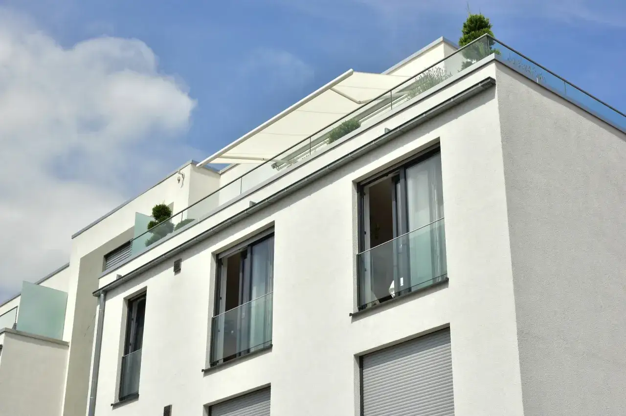 Nowoczesny budynek z białym tynkiem, szklanymi balustradami i wysuwanym markizą. Widoczny portfenetr z roślinnością na dachu.