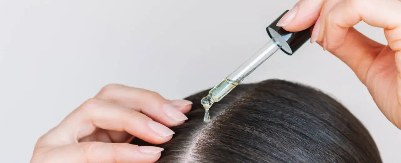 Eine Hand trägt mit einer Pipette Öl auf die Kopfhaut auf. Dieses Öl, oft Rizinusöl, wird für die Haarpflege verwendet.