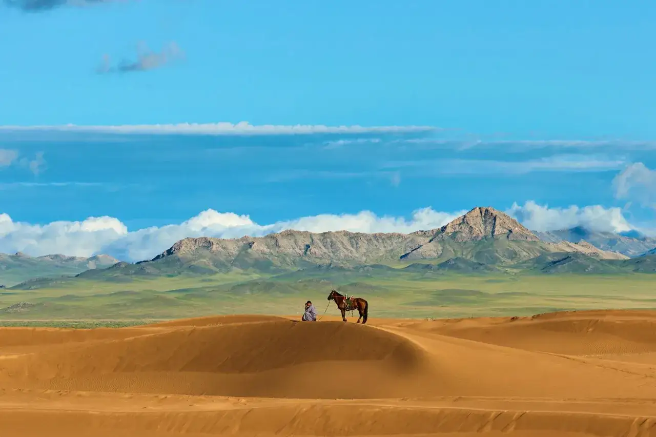Eine Person und ein Pferd ruhen auf einer Sandd&uuml;ne vor einer majest&auml;tischen Bergkette. Eine typische Mongolei Landschaft.