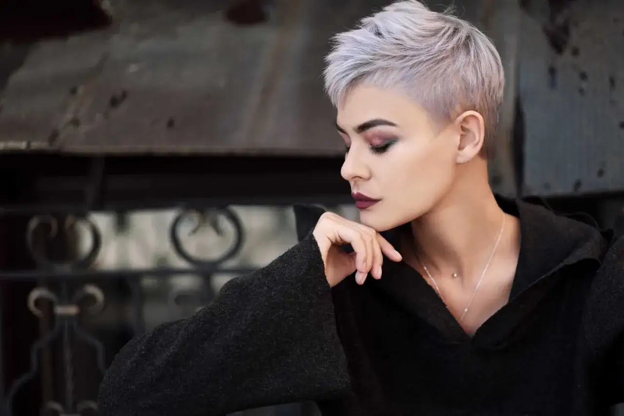 Mujer joven con pelo blanco corto y maquillaje dramático, apoyando la barbilla en la mano.