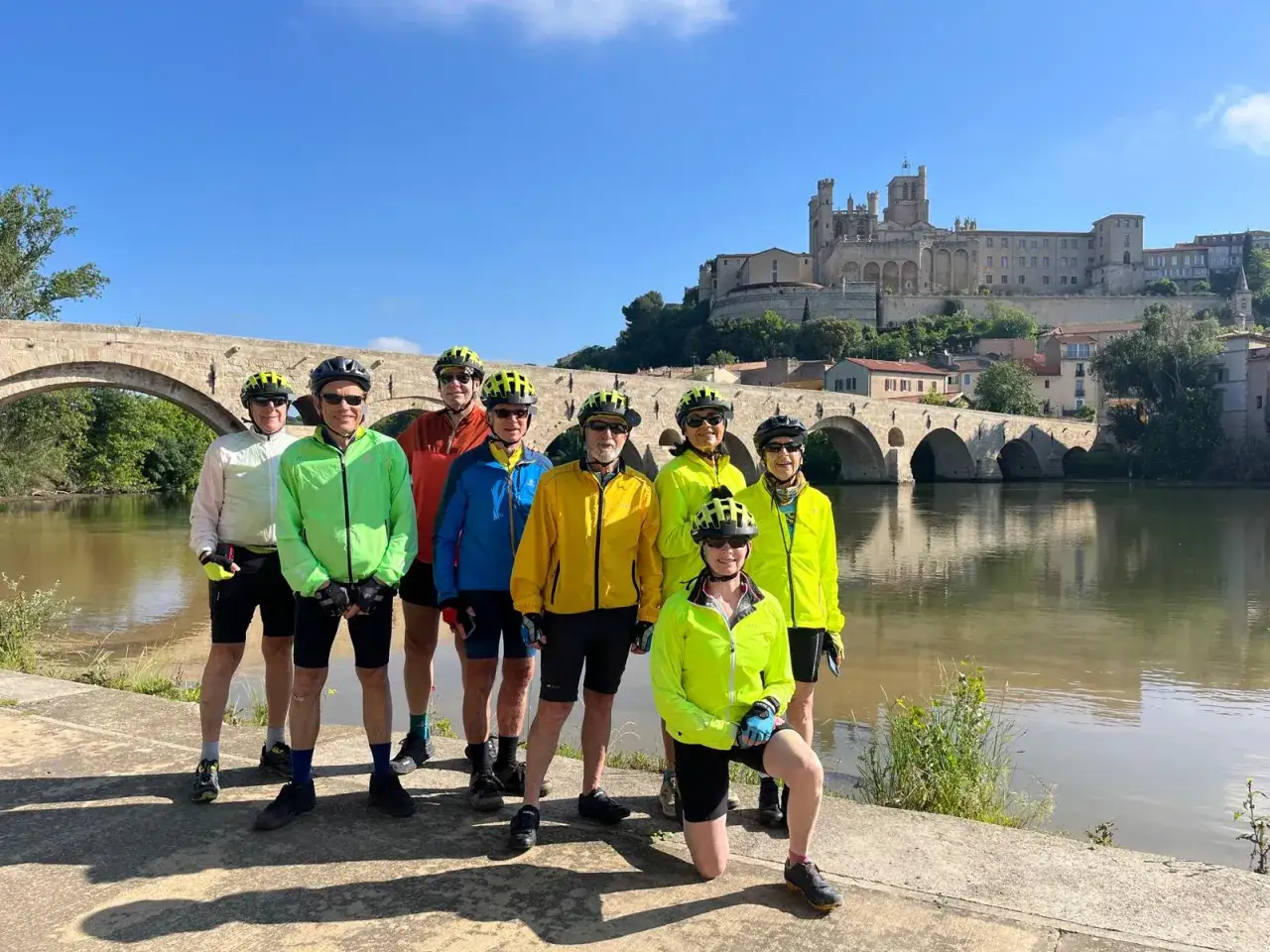 A group of cyclists on a canal du midi bike tour pose by a river with a historic bridge and cathedral in the background.