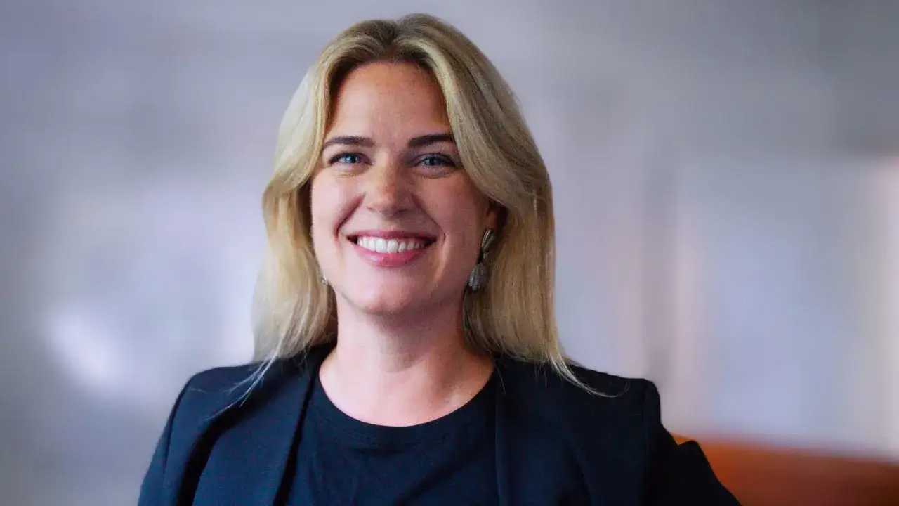 Amanda Oxell, en kvinna med blont hår och blå ögon, ler mot kameran. Hon bär en svart kavaj och en svart topp.