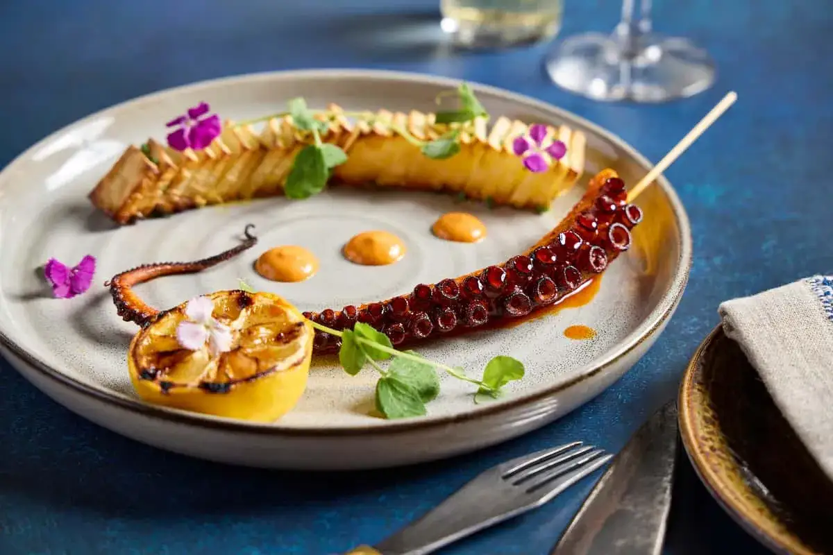 Delicioso pulpo a la plancha, servido con limón asado, flores moradas y una salsa cremosa.