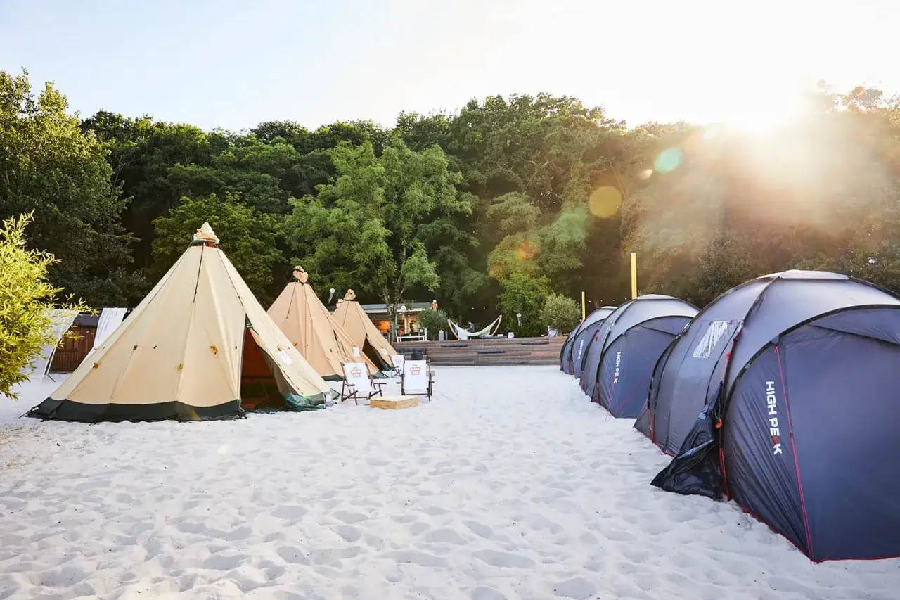 Ein **Summerjam Comfort Camp** mit Tipizelten und modernen Zelten am Strand, umgeben von Bäumen.