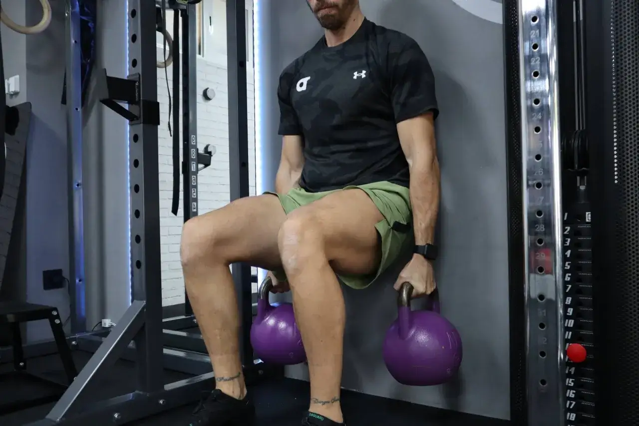 Hombre realizando sentadilla isom&eacute;trica en pared con pesas rusas. Una tabla de ejercicios isom&eacute;tricos es ideal para este entrenamiento.