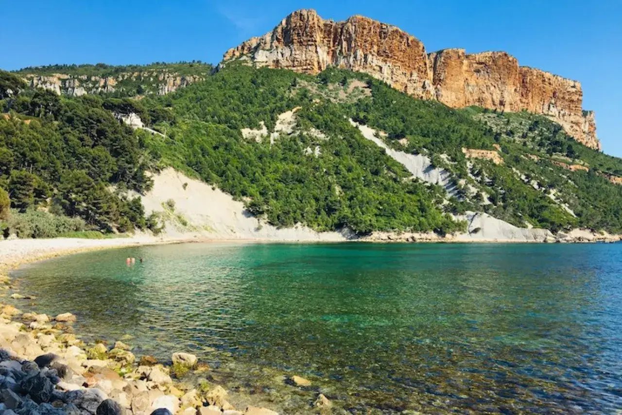 Plage rocheuse aux eaux turquoise sous le majestueux Cap Canaille. Deux baigneurs profitent de la quiétude.