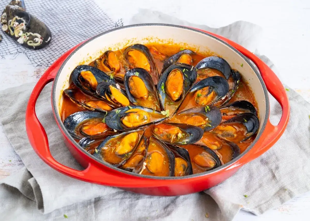 Mejillones a la marinera recién cocinados en una cazuela roja, con una salsa sabrosa y perejil picado.
