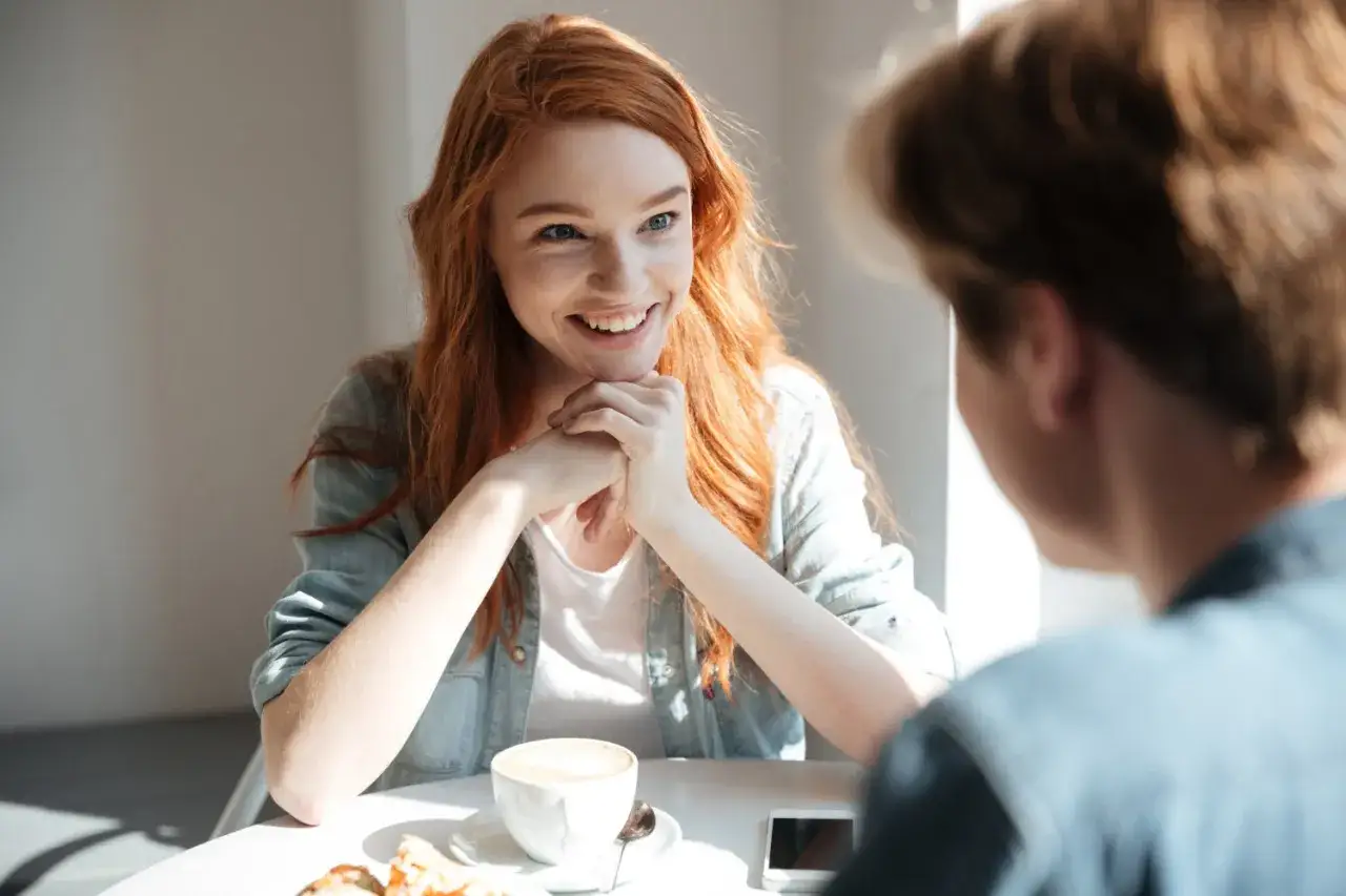 Uśmiechnięta rudowłosa dziewczyna rozmawia z chłopakiem przy kawie. Ich spojrzenia pełne są ciepła, świadcząc o pięknej miłości platonicznej.