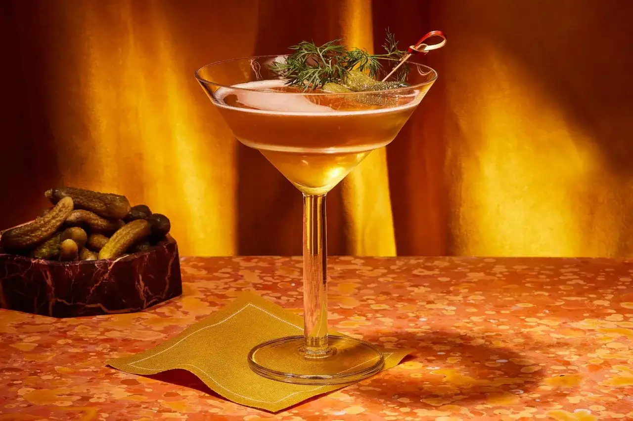 Un cocktail ambré, garni d'aneth et de cornichons, évoque un nom de cocktail féminin. À côté, un bol de cornichons et une serviette dorée.