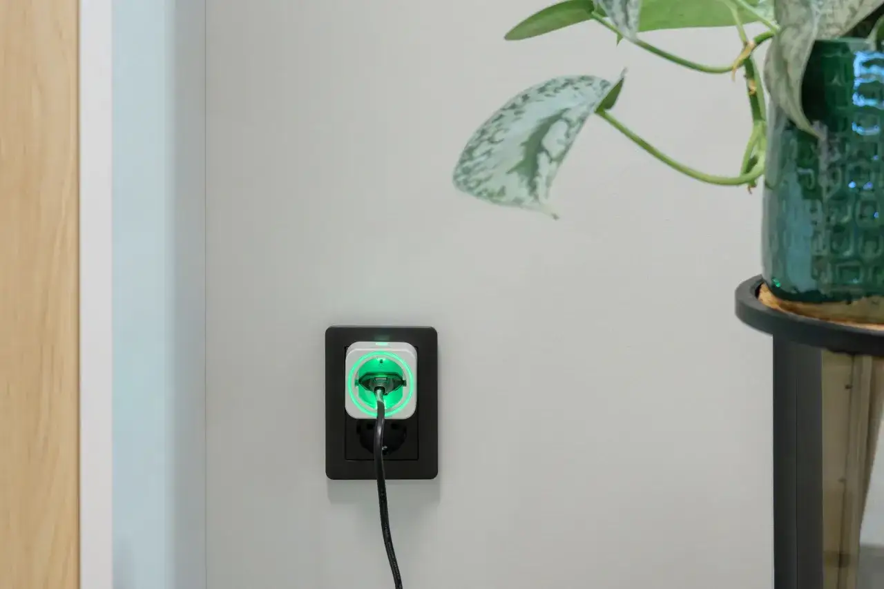 En vit smart kontakt med grön lysande ring sitter i ett svart vägguttag. Bredvid en grön kruka med en växt. En mekanisk timer kan styra detta.