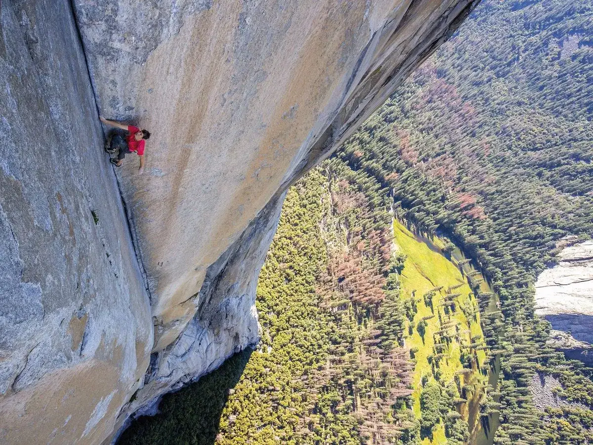 Ein Kletterer wagt sich im **free solo** Stil an einer riesigen Felswand empor, während unten ein grünes Tal mit Bäumen und einem Fluss liegt.