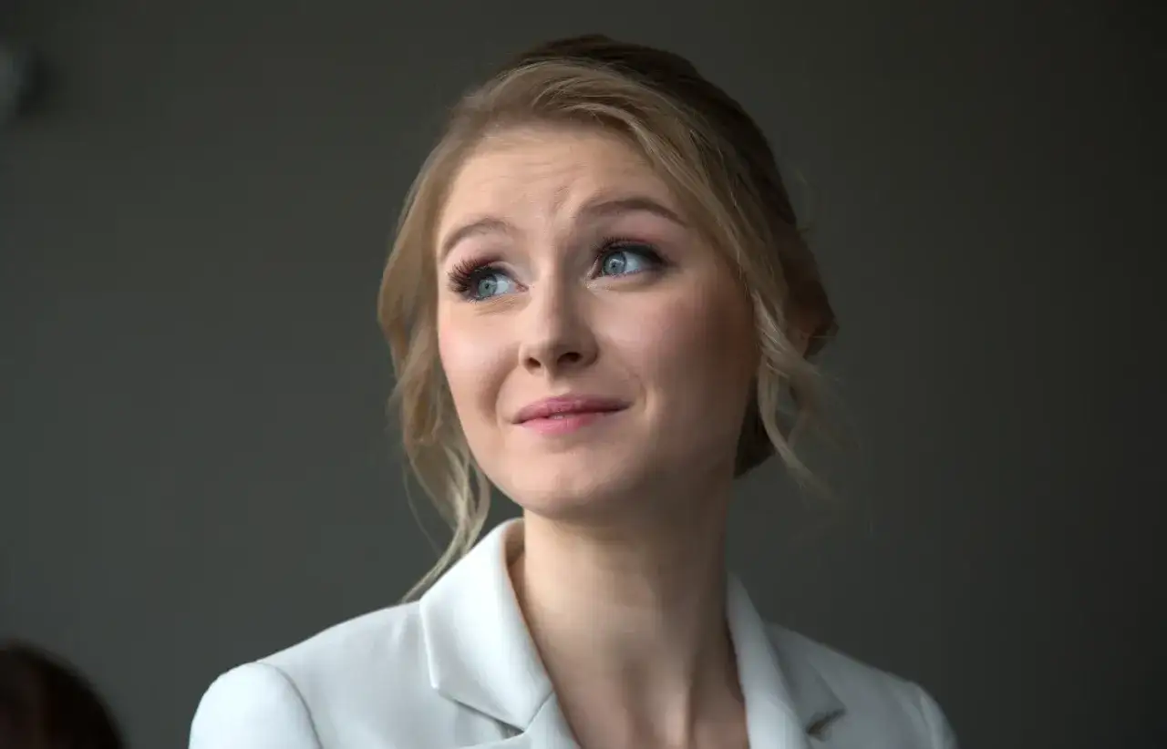 Portret młodej kobiety z blond włosami i niebieskimi oczami, w białej marynarce. Wiek Kingi Dudy jest trudny do określenia.