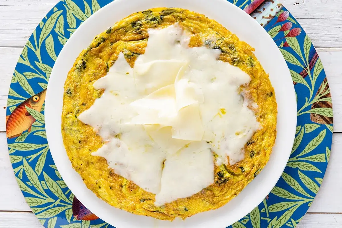 Frittata di bietole dorata e filante, servita su un piatto bianco con un sottobicchiere blu decorato con pesci e foglie.