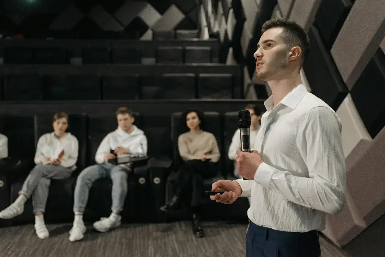 A young man confidently speaks into a microphone, finding his voice on stage as an audience watches.