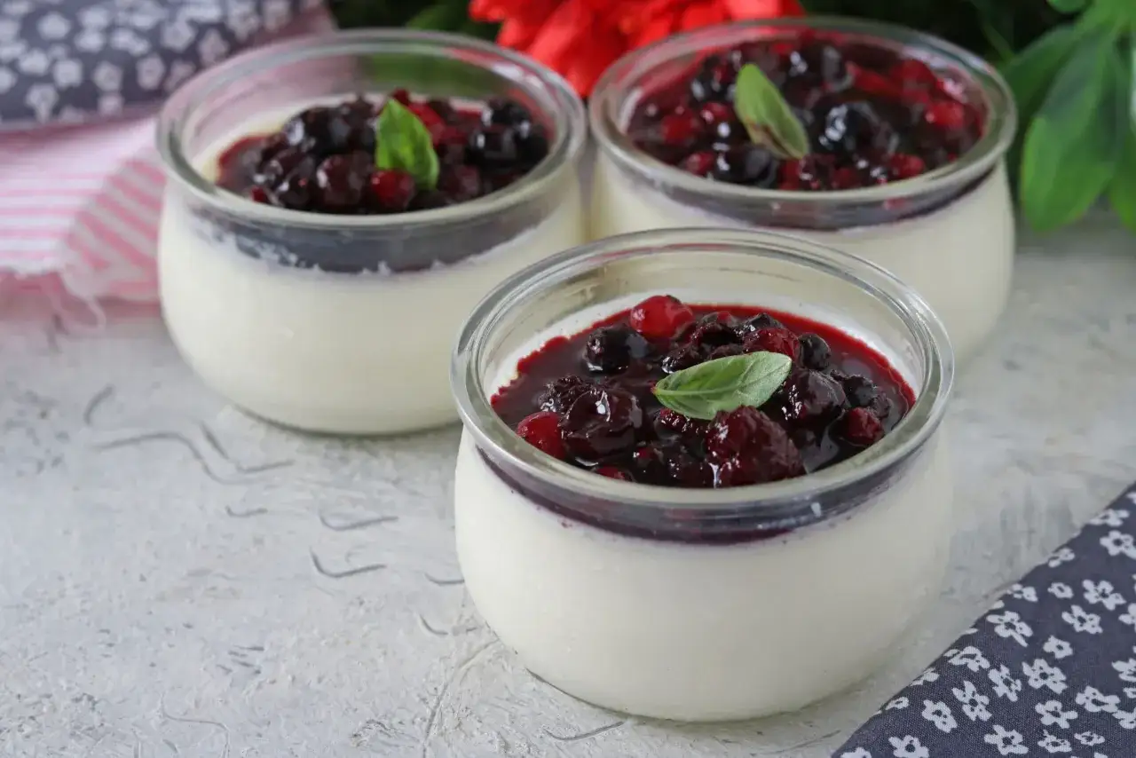 Tre deliziose panna cotta in vasetti di vetro, guarnite con frutti di bosco e una fogliolina di menta.