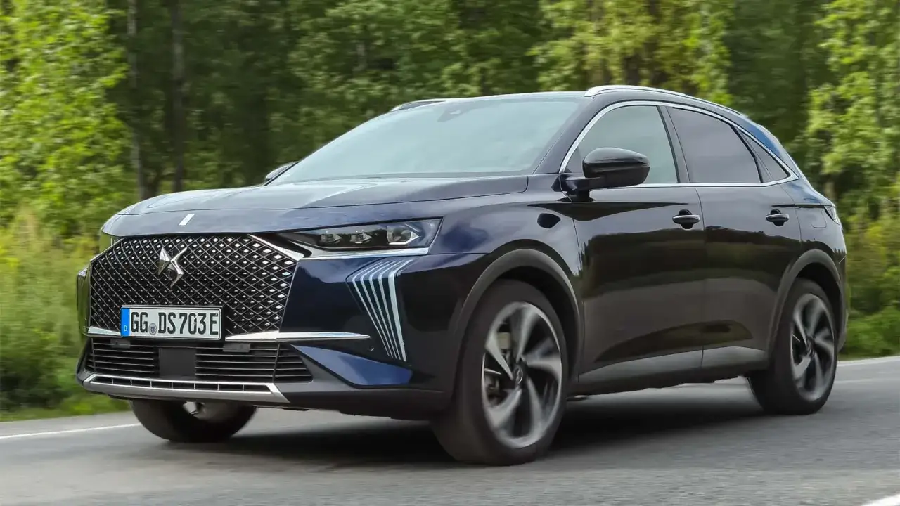 Ciemnoniebieski DS 7 Crossback E-Tense sunie po drodze, otoczony zielenią.