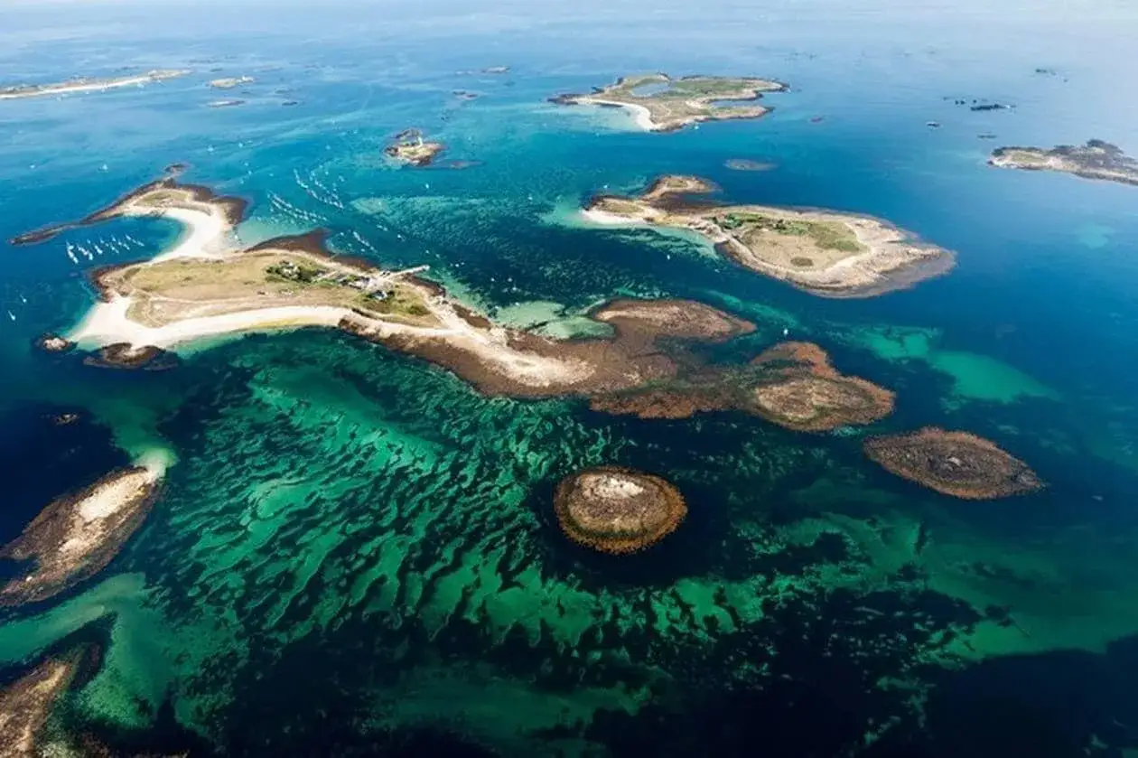 Vue aérienne d'un archipel breton aux eaux turquoise et vertes, parsemé d'îlots rocheux et de bancs de sable.