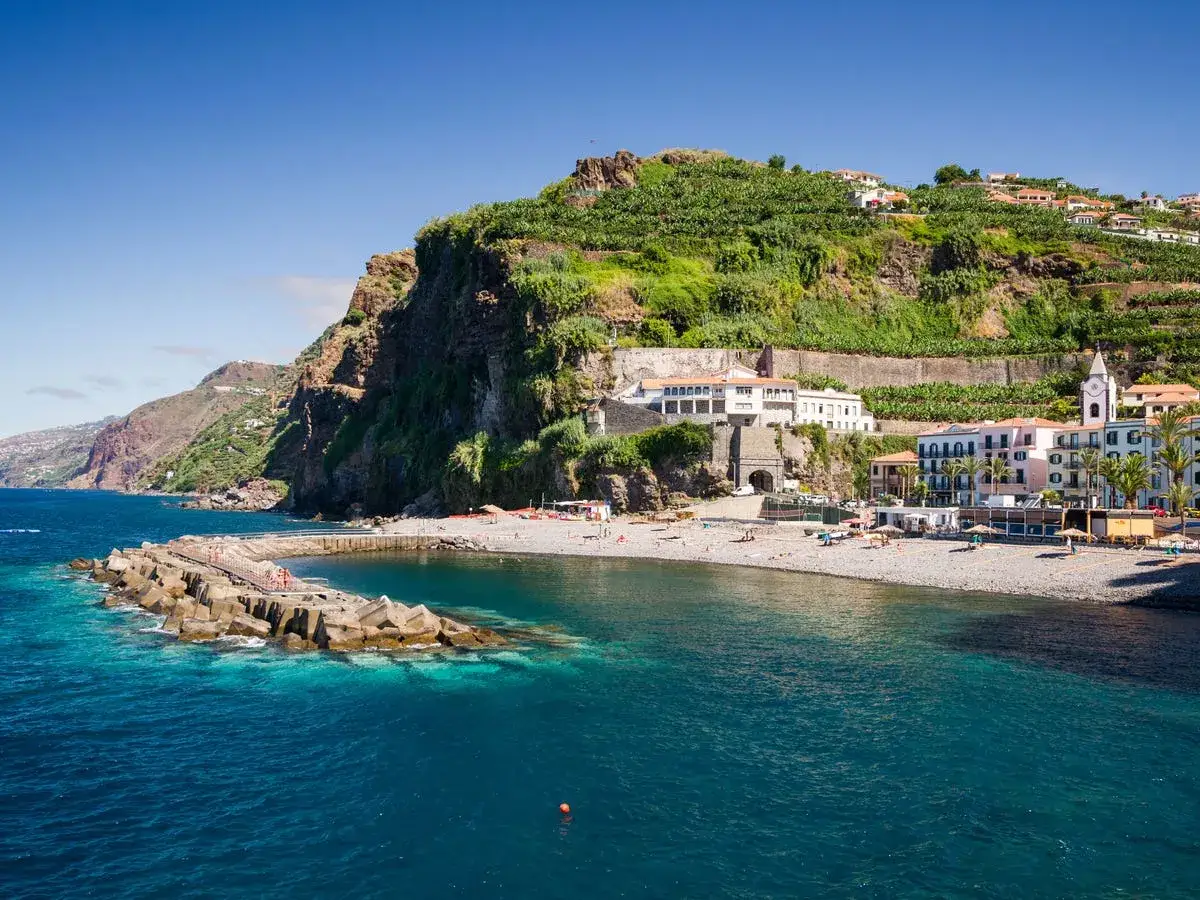 Costa rocosa con pueblo costero y playa de guijarros en las islas de Portugal. Palmeras y edificios blancos y rosas bajo colinas verdes.