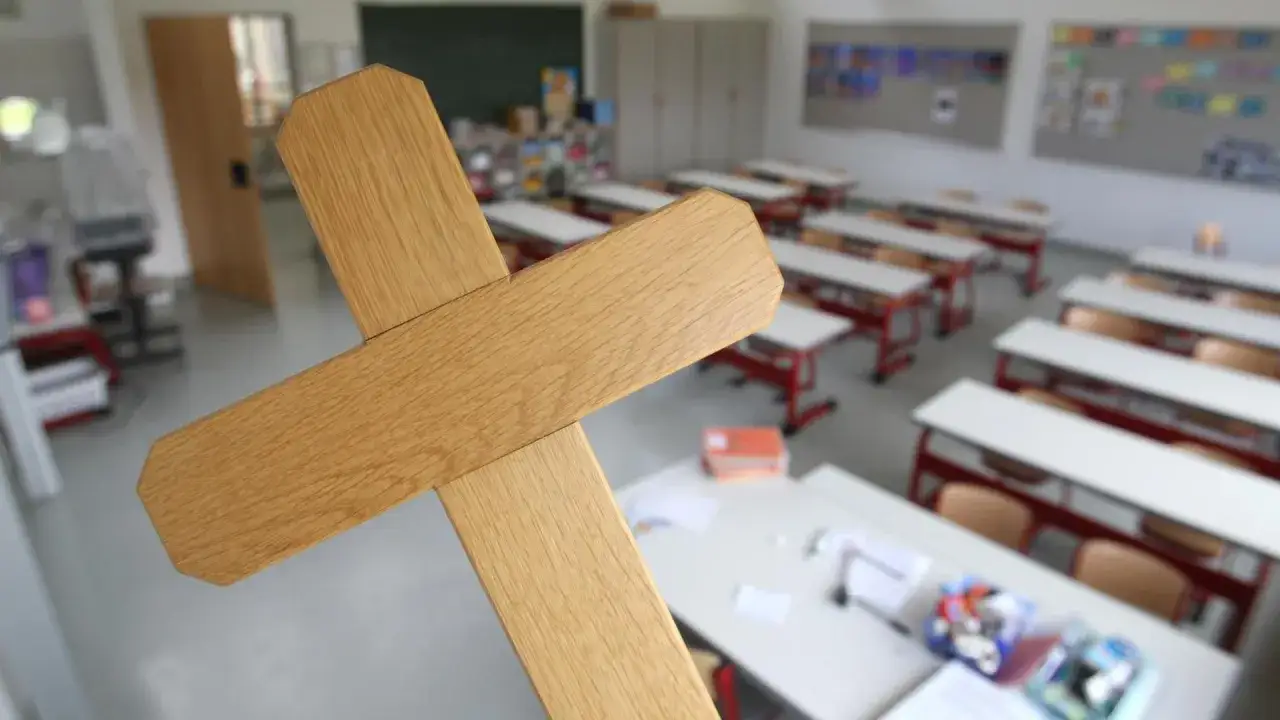 Holzkreuz vor einem leeren Klassenzimmer. Ein Symbol, das in säkularen Schulen Fragen aufwirft.